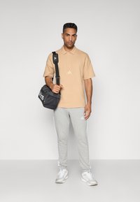 Beige textuur polo shirt met korte mouwen, grijze joggingbroek met witte strepen en een zwarte Adidas schoudertas. Witte sneakers maken de look compleet.
