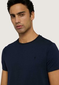 Námořnická modrá tričko s krátkým rukávem a kulatým výstřihem vyrobené z měkké bavlny. Obsahuje malé vyšívané logo na levé straně hrudi. Krátké rukávy, hladká textura.