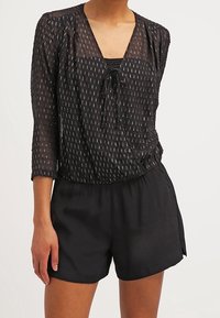 Top negro transparente con mangas 3/4, que presenta líneas verticales blancas, un escote en V y un lazo de ajuste en la parte delantera, combinado con pantalones cortos negros.
