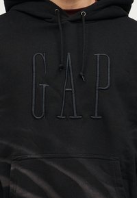 Μαύρο φούτερ με κουκούλα, με μεγάλο κεντητό λογότυπο GAP στο μπροστινό μέρος και μαύρα κορδόνια που κρέμονται από τη γωνία της κουκούλας.