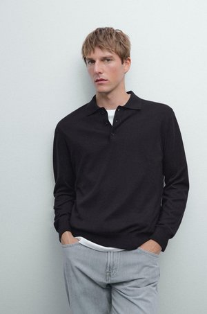 Young man in black long-sleeve polo shirt layered over a white top, wearing light gray jeans, standing with hands in pockets against plain wall.