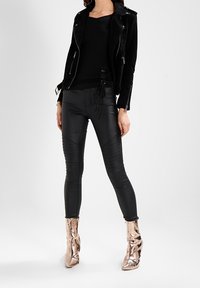 Zwarte suède motorjack, aansluitend zwart topje, glanzende zwarte legging met textuurdetails, en metallic roségouden enkellaarsjes.