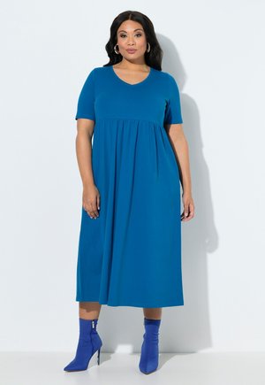 Frau mit langen dunklen Haaren, die ein blaues Midikleid mit kurzen Ärmeln und passende blaue Stiefeletten mit Absatz trägt, steht vor einem weißen Hintergrund.