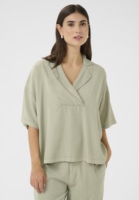 Gaiši zaļa, īsām piedurknēm blūze ar v-veida kakla izgriezumu, atbrīvota piegriezuma un viegla auduma. Iezīmējas ar smalku spīdumu un sānu kabatām.