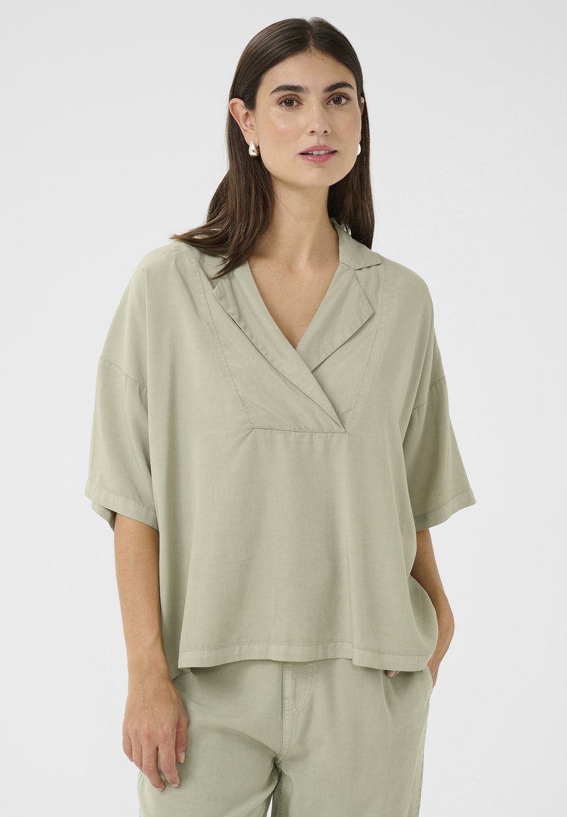 Gaiši zaļa, īsām piedurknēm blūze ar v-veida kakla izgriezumu, atbrīvota piegriezuma un viegla auduma. Iezīmējas ar smalku spīdumu un sānu kabatām.