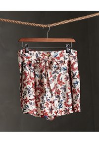 Pantalones cortos con estampado floral en tela crema, con flores rojas y azules, cintura con lazo y diseño plisado. Cuelgan de una percha de madera.