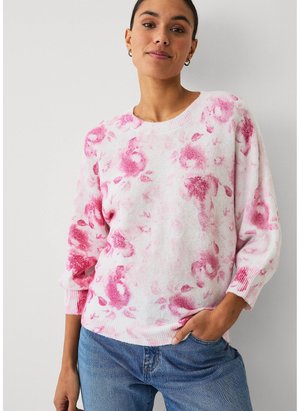 Vrouw met een witte trui met roze bloemenpatronen en een blauwe spijkerbroek, staand met één hand in haar zak tegen een effen achtergrond.