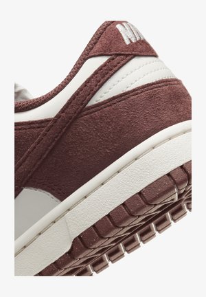 Primer plano del talón de una zapatilla de gamuza burdeos y cuero blanco con suela texturizada y detalles visibles de costura.