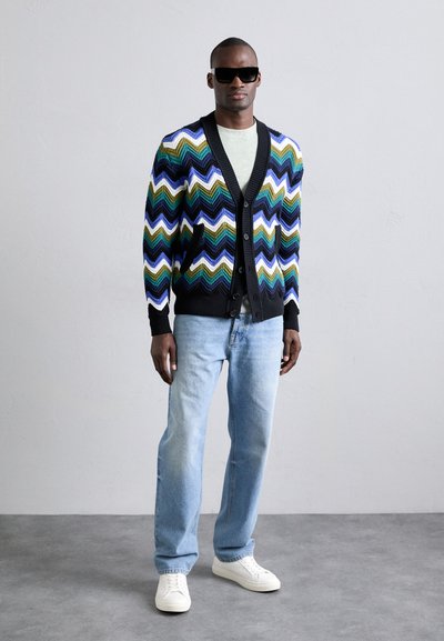 Cardigan colorat cu un model în zigzag în negru, verde, albastru și alb. Purtați peste un tricou gri deschis, asortat cu blugi albaștri deschiși și adidași albi.