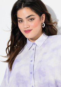 Camicia blu viola chiaro con un motivo tie-dye, morbida al tatto e con colletto classico, caratterizzata da bottoni viola. Orecchini a cerchio argentati visibili.