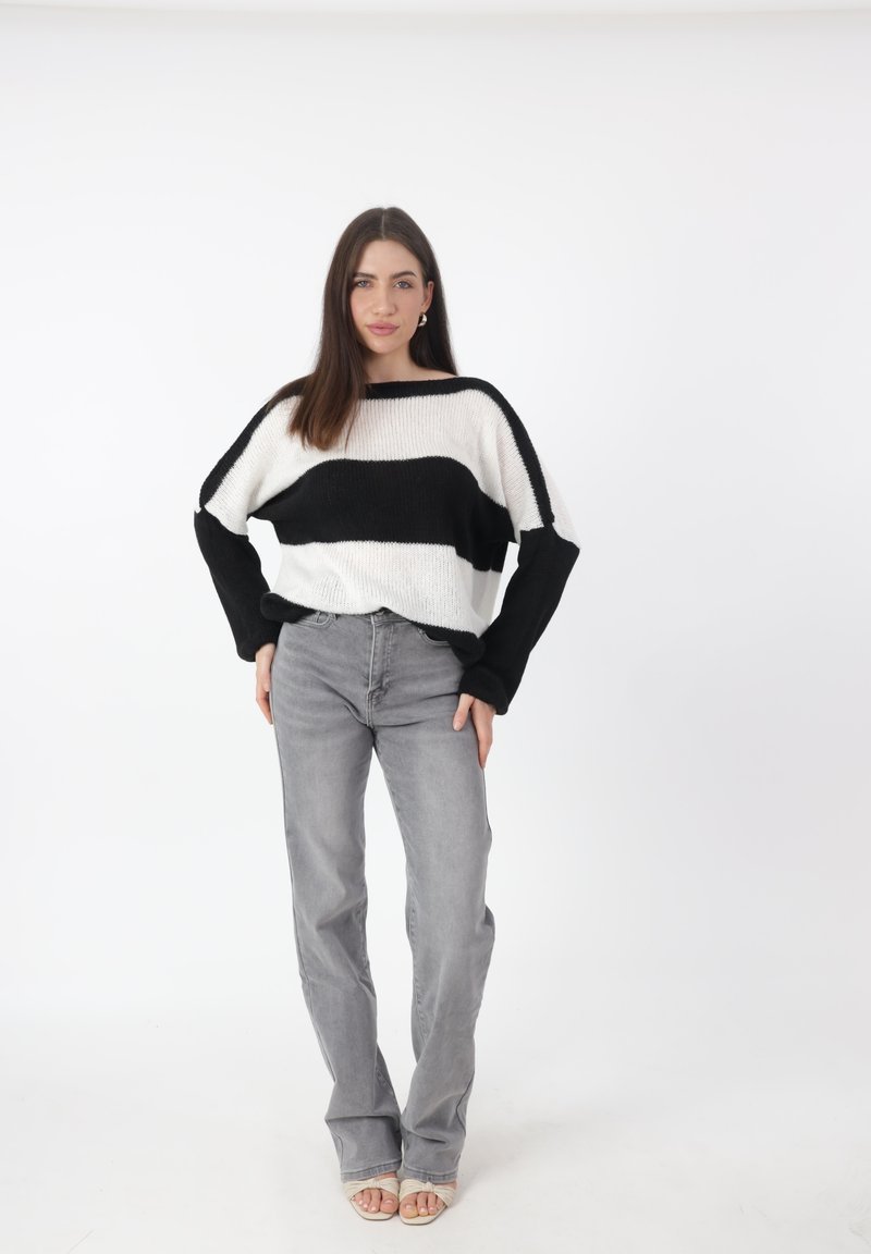 Maglione oversize a righe bianche e nere con maniche ampie, abbinato a jeans grigi chiari a zampa d'elefante e sandali beige su uno sfondo neutro.