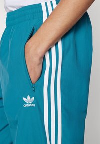 Pantaloni da track teal di Adidas con strisce bianche, vita elasticizzata, tasca laterale con zip, e dettaglio del logo sul davanti. Tessuto testurizzato.