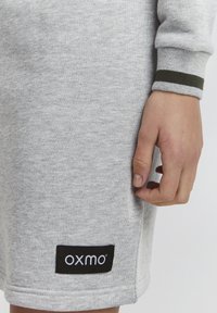 Sivi dukserica od tkanine s teksturiranim finišom, koja sadrži crnu oznaku s "OXMO" i zelene prugaste detalje na manžetama.