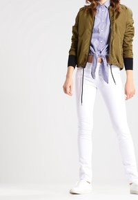 Chaqueta bomber verde oliva con puños negros sobre una camisa de rayas azul y blanca anudada en la cintura, combinada con jeans skiny blancos y zapatillas blancas.