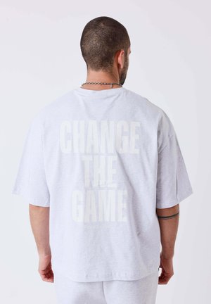 Homme aux cheveux courts avec un tatouage sur le bras portant un T-shirt gris clair oversize avec le texte "CHANGE THE GAME" au dos.