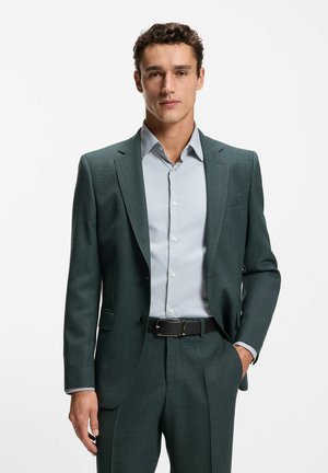 Man draagt een donkergroen pak met een lichtblauw overhemd en een zwarte riem, staat met één hand in zijn zak.