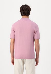 Mand set fra ryggen iført en lysegrå kortærmet polo-shirt og off-white bukser foran en ensfarvet hvid baggrund.