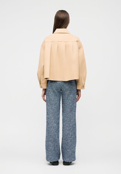 Γυναίκα όρθια, φοράει μπεζ cropped παλτό με πιέτες στην πλάτη και μπλε υφασμάτινο παντελόνι με φαρδιά μπατζάκια, μαύρα παπούτσια, με την πλάτη προς τα εμπρός.