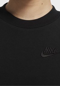 Svart sweatshirt med ribbad krage och en svart broderad Nike-logotyp på bröstet. Materialet är slätt med en strukturerad yta.