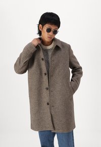 Cappotto marrone con trama e colletto, dotato di chiusura a bottoni, due tasche laterali, indossato sopra un maglione a motivo, abbinato a occhiali da sole.