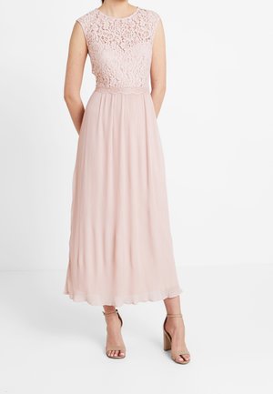 Kvinde iført ærmeløs, lyserød blondekjole med plisseret nederdel og beige højhælede sandaler, stående mod en hvid baggrund.