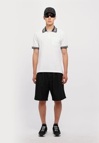 Biele polo tričko s čiernymi akcentmi, zodpovedajúce čierne šortky a čierna čiapka. Slnečné okuliare dokončujú športový outfit. Jednoduchý, moderný dizajn.