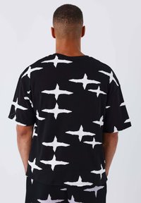 T-shirt en coton noir avec un motif abstrait de croix blanche. Coupe décontractée avec des manches courtes et un col rond, design uni à l'arrière.