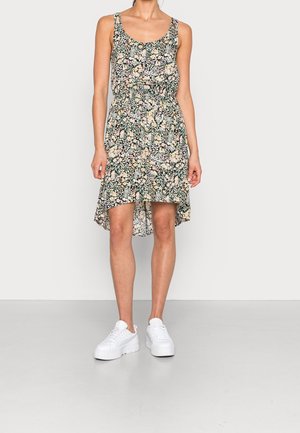 Femme portant une robe sans manches à motifs floraux avec un ourlet asymétrique et des baskets blanches à plateforme, debout devant un fond clair uni.