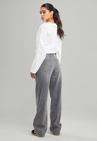 Jean gris à jambes larges avec poches arrière visibles et accents brodés, associé à un haut à manches longues blanc. Texture de tissu lisse.