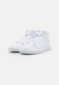 Pár bílých vysokých tenisek se šňůrkami, perforovanou špičkou a logem swoosh na boku, vystavených na jednoduchém bílém pozadí.