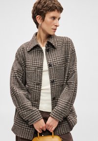 Brūnas krekliņš ar pārklājumu, ar apkakli, lielām pogām un strukturētu audumu, apvienots ar krēmu toni augšu un sinepju krāsas rokassomu.