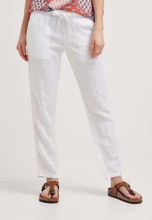 Vrouw die witte linnen broek met trekkoord en bruine sandalen draagt, staand tegen een eenvoudige lichte achtergrond.