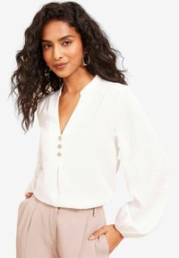Blusa bianca a maniche lunghe con scollatura a V e tre bottoni dorati. Tessuto leggero e strutturato; indossata con pantaloni beige chiaro.
