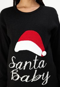 Black sweater featuring a festive red and white Santa hat design. The text "Santa Baby" is embroidered in white cursive. Soft texture.