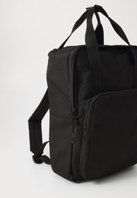 Czarny plecak materiałowy z regulowanymi paskami, zamknięciem na zamek błyskawiczny w głównym pionowym komorze oraz kieszenią z przodu. Teksturowana powierzchnia i prosty design.