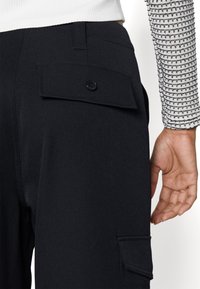 Personne portant un pantalon noir avec un dos boutonné et des poches latérales à rabat, ainsi qu'une chemise blanche à manches longues avec des rayures noires horizontales.