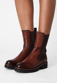 Bottes de cheville en cuir marron avec des panneaux latéraux élastiques noirs et de épaisses semelles crantées noires, portées sur des jambes nues sur fond blanc.