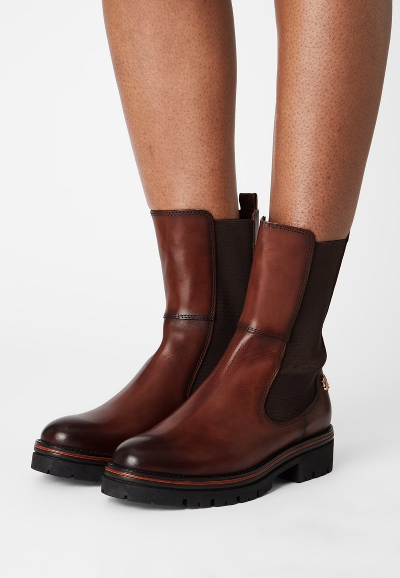 Bottes de cheville en cuir marron avec des panneaux latéraux élastiques noirs et de épaisses semelles crantées noires, portées sur des jambes nues sur fond blanc.
