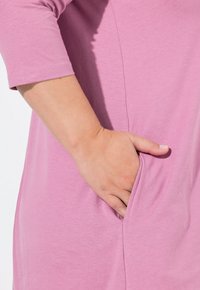 Helles rosa Kleid aus weichem Stoff, mit Dreiviertelärmeln und einer seitlichen Tasche auf Taillenhöhe, glatte Textur und schlichtes Design.