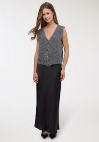 Gilet grigio lavorato a maglia con scollo a V, quattro bottoni decorativi, abbinato a una lunga gonna di raso nera e scarpe nere con punta.