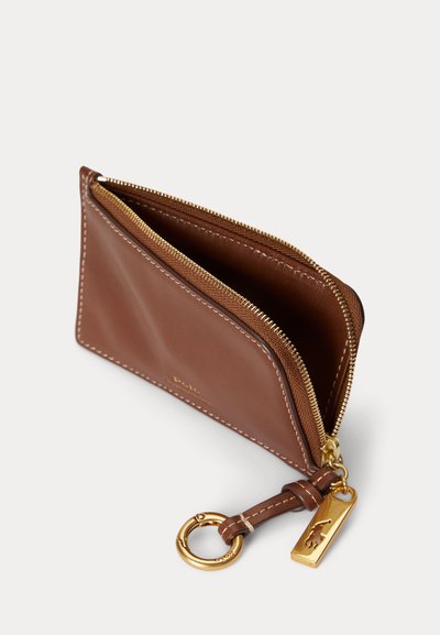 Καφέ δερμάτινη πορτοφολιέρα με φερμουάρ για κέρματα, χρυσό μεταλλικό μπρελόκ και ετικέτα με λογότυπο, εμφανίζεται μερικώς ανοιχτή σε λευκό φόντο.