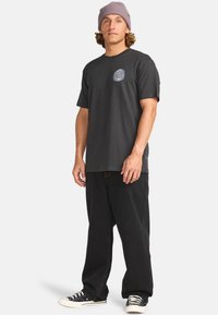 Svart bomull t-skjorte med korte ermer, med et grafisk logo på brystet. Kombinert med løse svarte bukser og hvite joggesko.