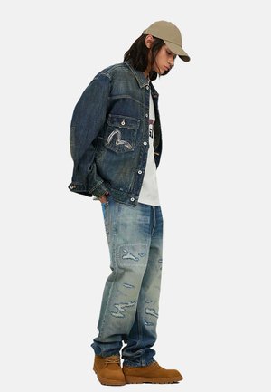 Giovane che indossa un berretto color camoscio, una giacca di jeans oversize, jeans larghi strappati, una t-shirt bianca con grafica e stivali marroni con lacci, guarda verso il basso.