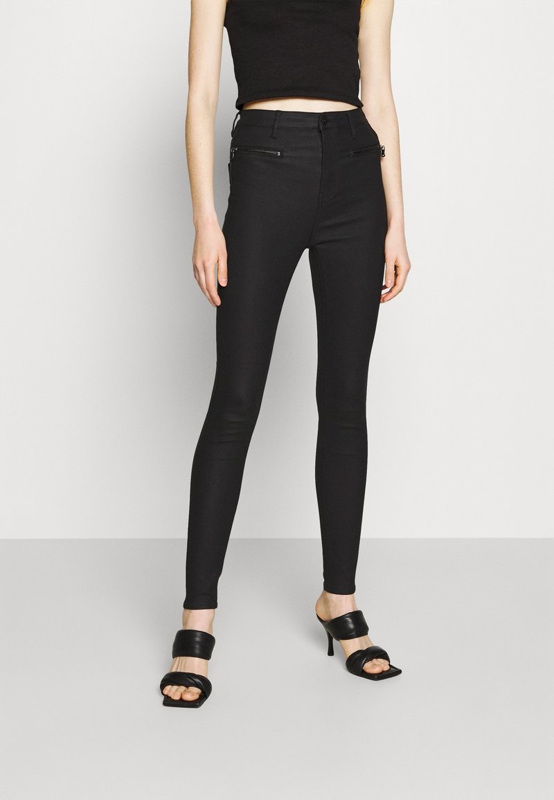 Jean skinny noir taille haute, avec des poches zippées à l'avant, associés à des sandales à talons noires, présentés sur un fond uni.