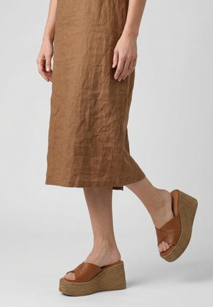 Person trägt einen braunen Leinenrock und hellbraune Ledersandalen mit Keilabsatz und geflochtenen Plateausohlen und steht mit einem leicht angehobenen Fuß.