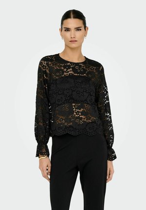 Mujer vistiendo una blusa de encaje negro con flores y manga larga, con dobladillo con forma de ondas, y pantalones negros, de pie contra un fondo claro y liso.