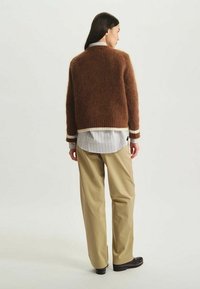 Pull en tricot marron avec des bords blancs, ayant un design carré. Assorti à une chemise rayée et un pantalon large beige.