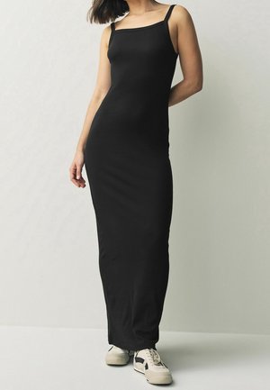 Femme portant une longue robe noire ajustée sans manches, associée à des baskets blanches et noires, debout devant un fond clair uni.