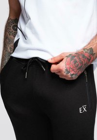 Czarne dresy z regulowanym paskiem na sznurku, kieszeniami na zamek i białym logo "EX". Charakteryzują się gładką teksturą materiału i dopasowanym krojem.