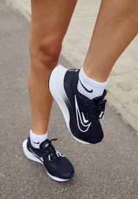 Zapatillas deportivas negras con acentos blancos, parte superior de malla y suela gruesa. Llevadas con calcetines blancos hasta la mitad en un fondo de pavimento.